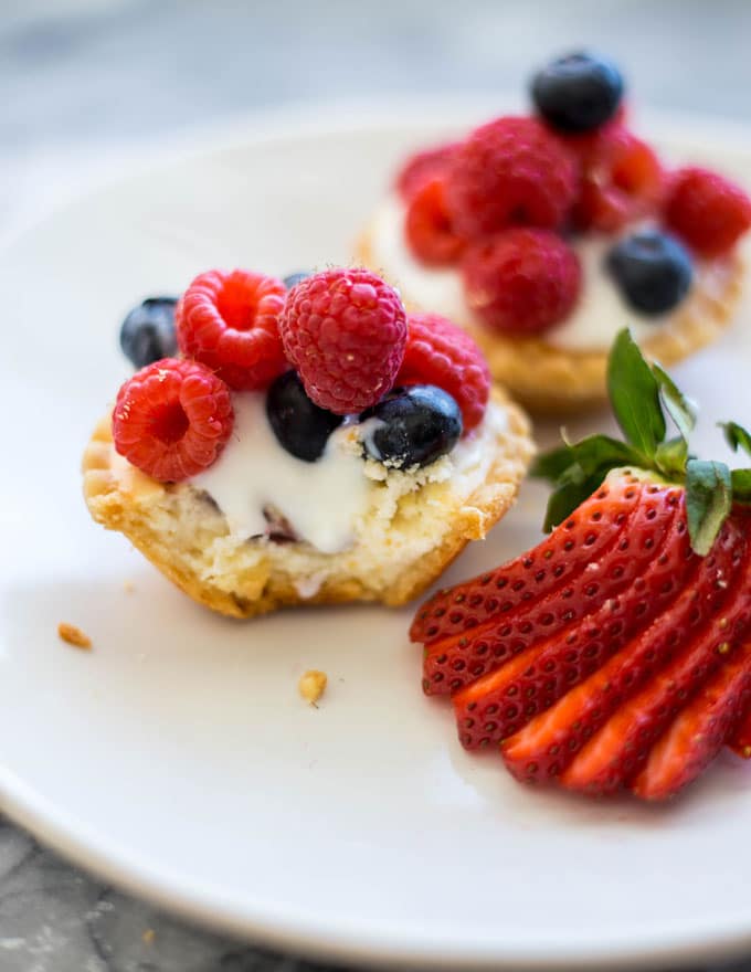a mini tart with a bite taken out topped with yogurt, raspberries, and blueberries on a plate with a strawbery