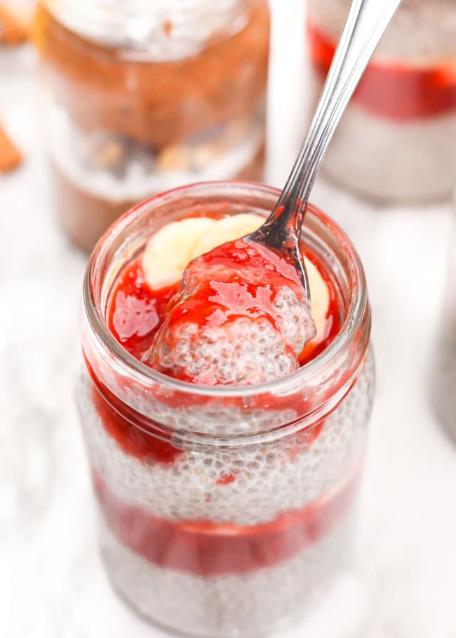 a spoon scooping chia pudding out of a jar with strawberry sauce and bananas