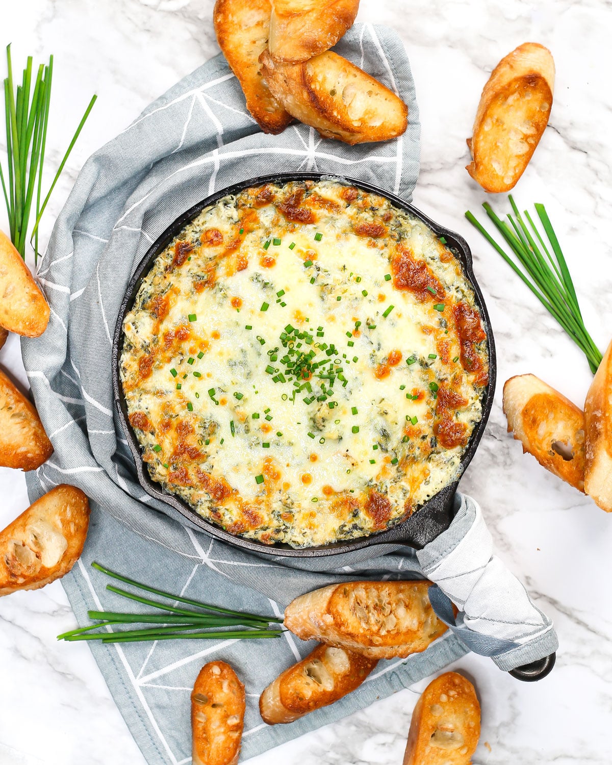 a skillet of spinach artichoke dip surrounded by crostini and chives