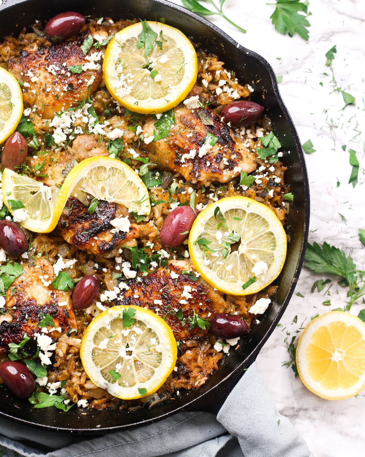 mediterranean chicken and rice in a skillet topped with lemons, olives, feta, and herbs.